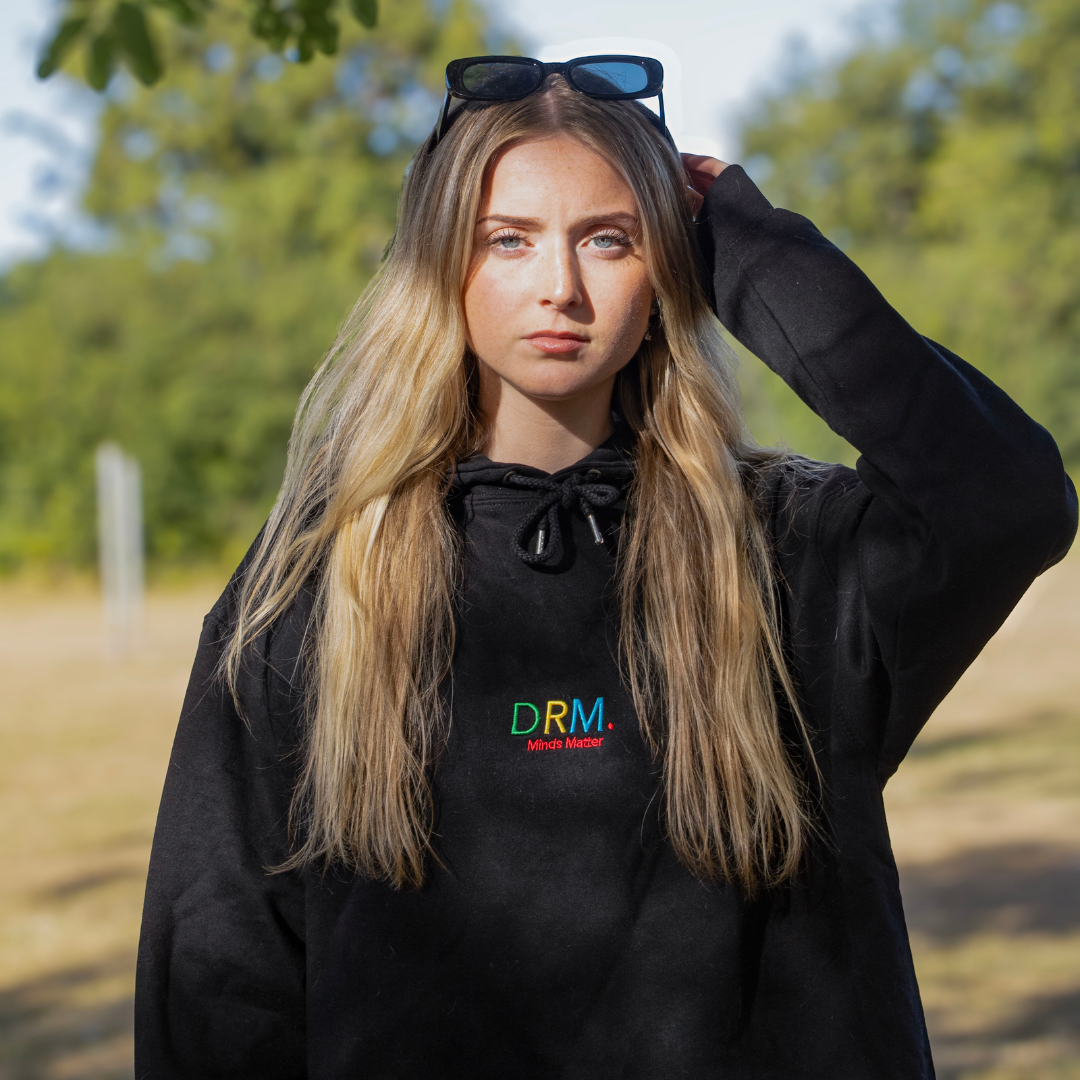 Person wearing a black hoodie with 'DRM' logo embroidered in multicoloured text standing outdoors
